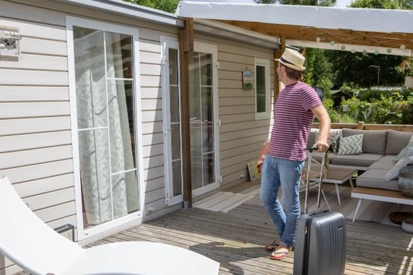 Homem arrastando uma mala de viagem sobre o deck de madeira em frente à casa, com porta de vidro de correr e área externa com sofá sob toldo, em dia de sol.