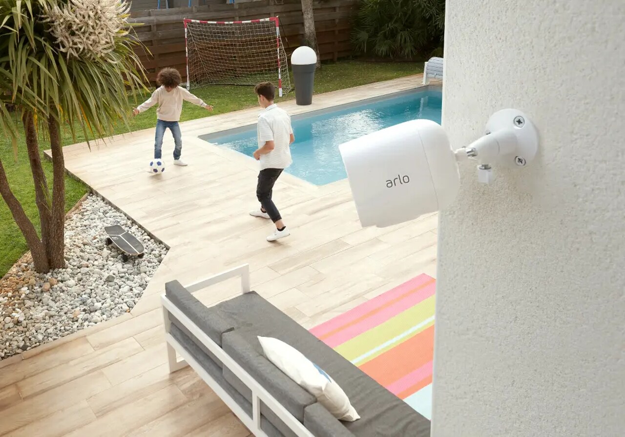 Crianças brincando em um quintal com piscina, gramado verde, árvore, rede de futebol e mobiliário externo.