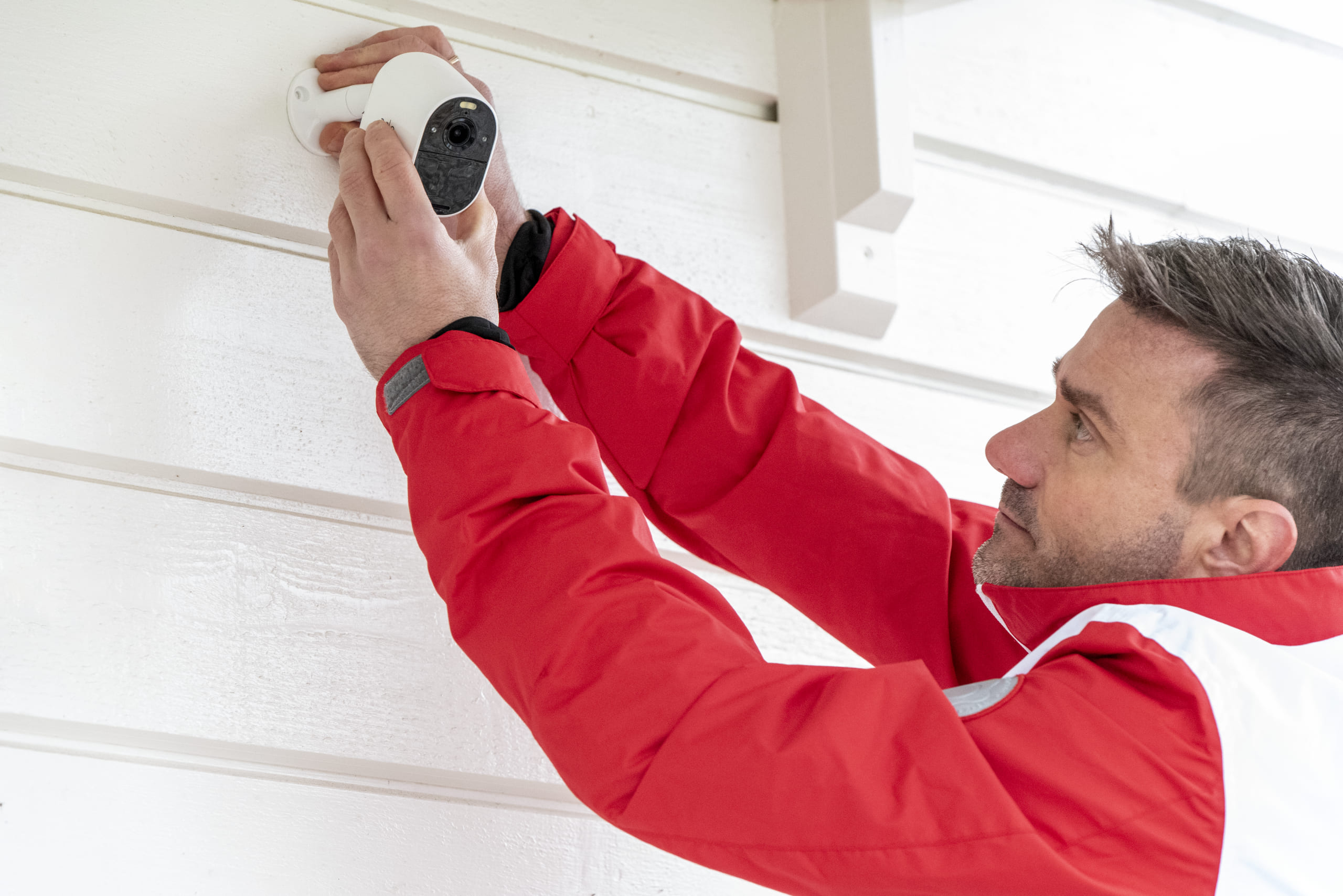 Profissional instalando câmera de segurança na parede de uma casa, vestindo jaqueta vermelha, focado na instalação para garantir a segurança.