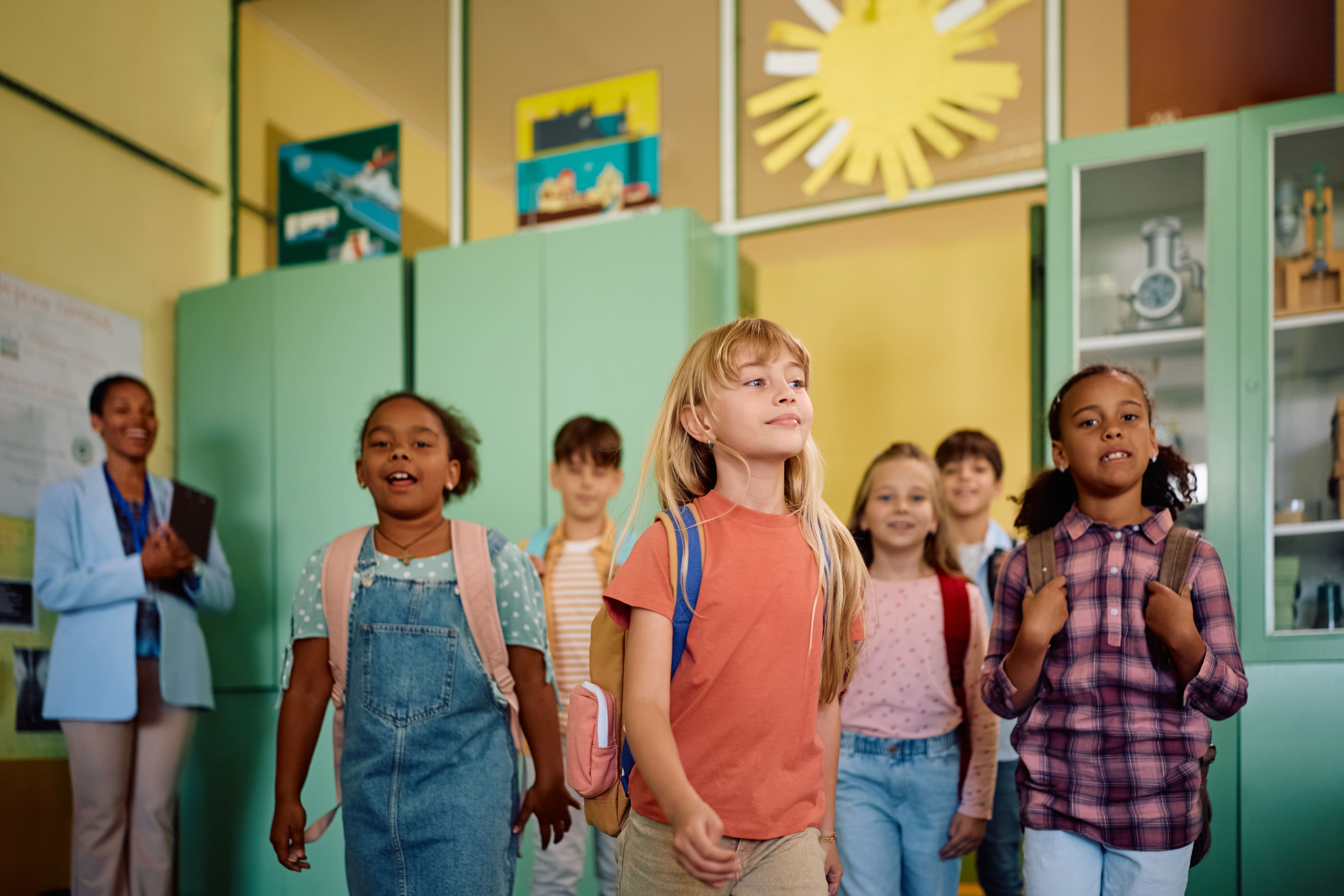 Crianças felizes e animadas entrando na escola, exibindo backpacks coloridos em um ambiente escolar com ambientes decorados e prateleiras com objetos didáticos.