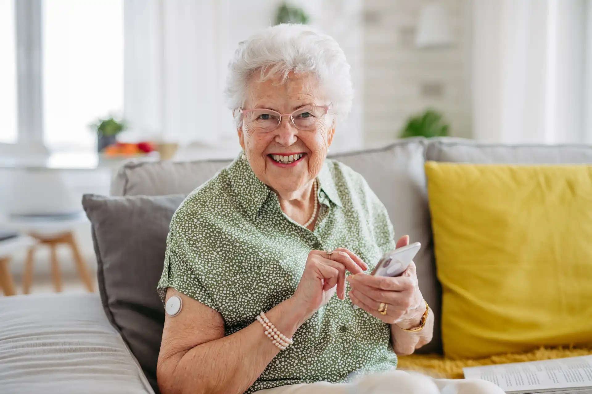 Mulher idosa sorridente usando aparelho de medição na braço enquanto usa celular em casa, representando saúde e bem-estar na terceira idade.