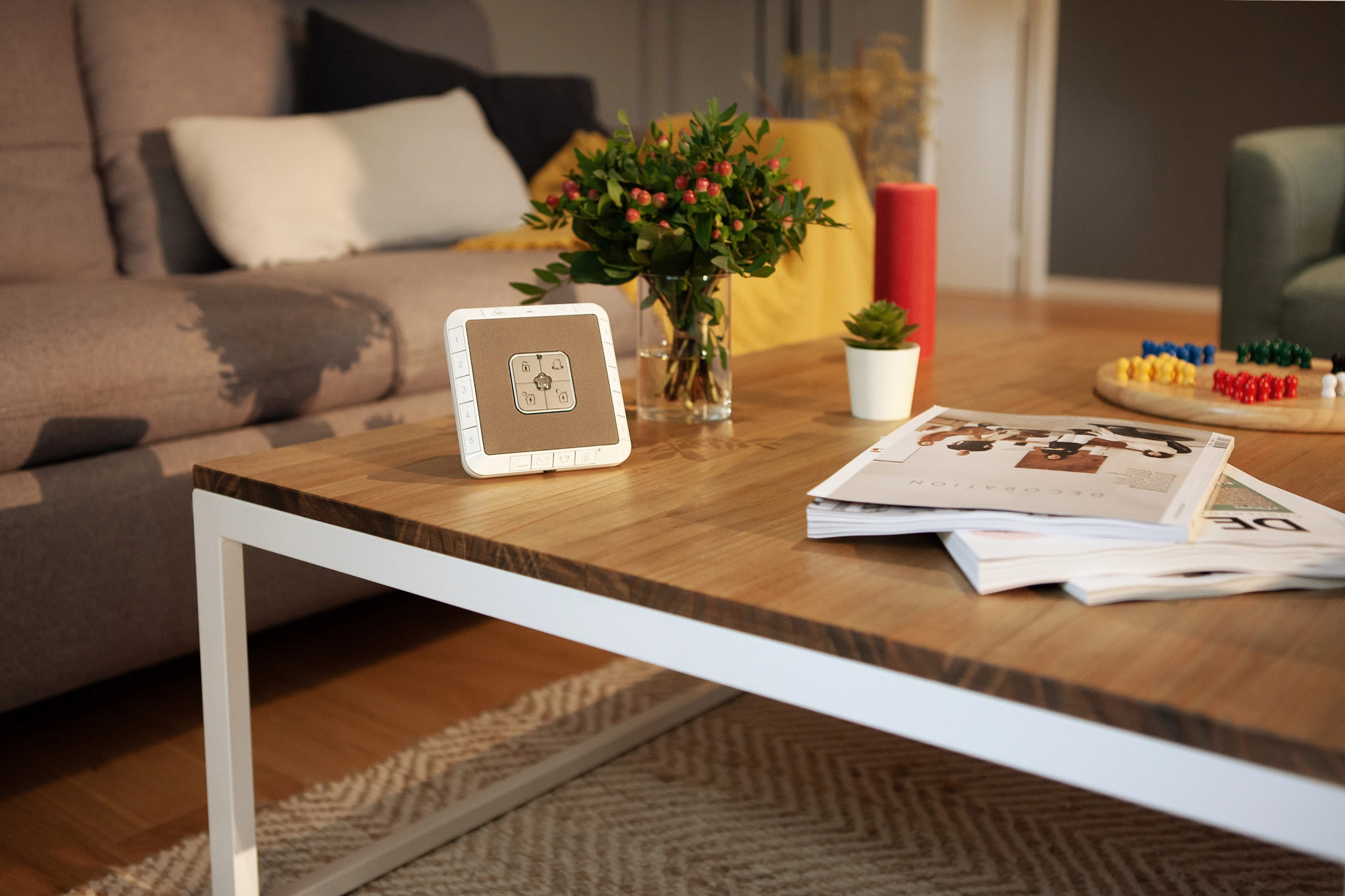 Imagem de uma sala de estar moderna com uma mesa de madeira com a central de alarmes Verisure, revistas, plantas e objetos decorativos ao redor.