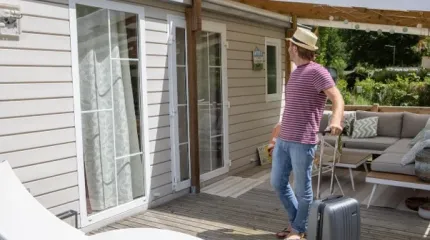 Homem arrastando uma mala de viagem sobre o deck de madeira em frente à casa, com porta de vidro de correr e área externa com sofá sob toldo, em dia de sol.