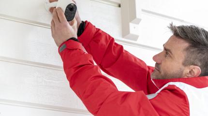 Profissional instalando câmera de segurança na parede de uma casa, vestindo jaqueta vermelha, focado na instalação para garantir a segurança.