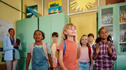 Crianças felizes e animadas entrando na escola, exibindo backpacks coloridos em um ambiente escolar com ambientes decorados e prateleiras com objetos didáticos.