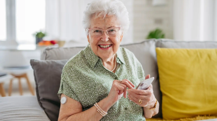 Mulher idosa sorridente usando aparelho de medição na braço enquanto usa celular em casa, representando saúde e bem-estar na terceira idade.
