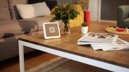 Imagem de uma sala de estar moderna com uma mesa de madeira com a central de alarmes Verisure, revistas, plantas e objetos decorativos ao redor.
