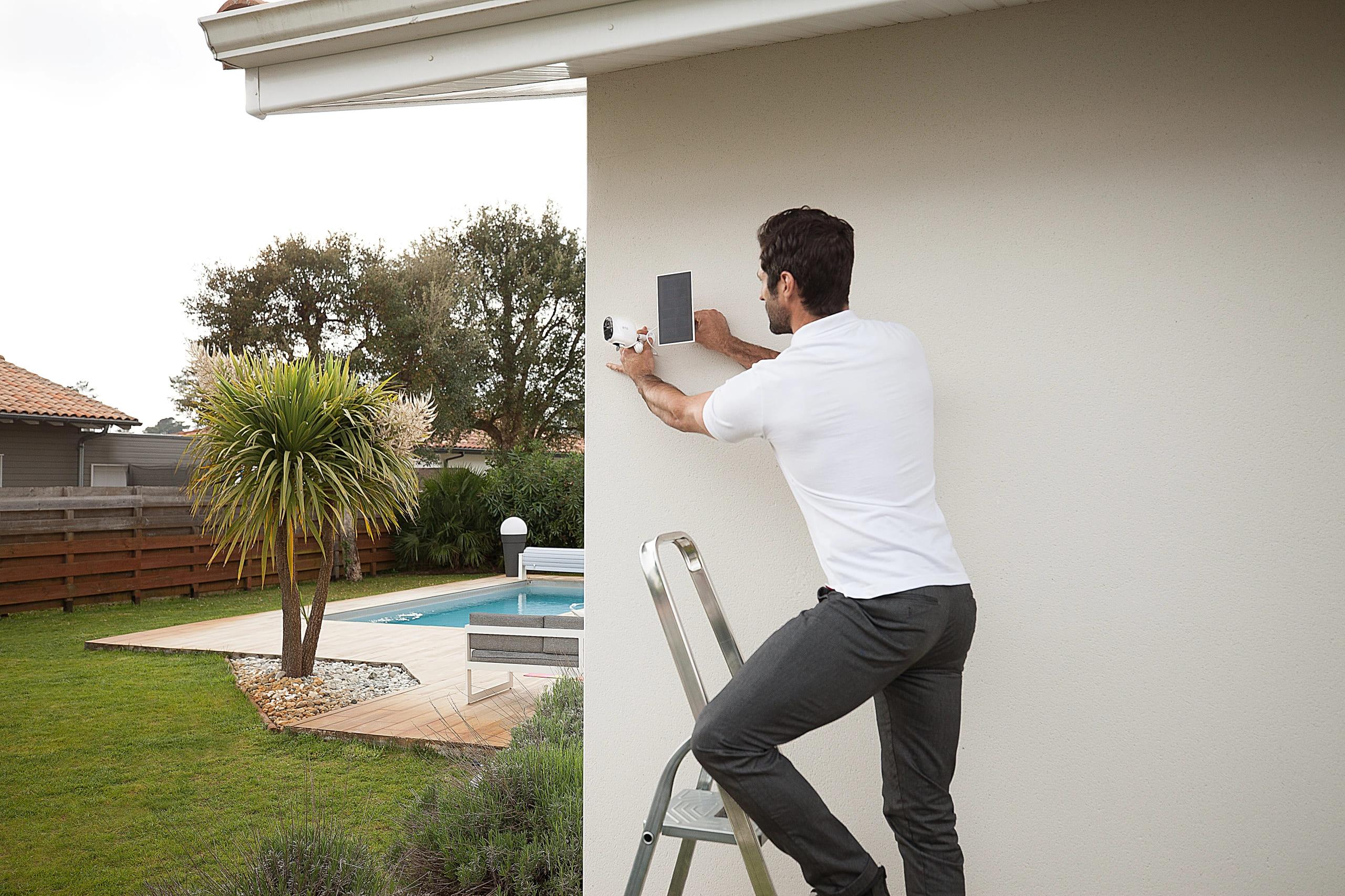 Homem instalando uma câmera de segurança na parede da casa no quintal, com piscina e jardim ao fundo, durante o dia.