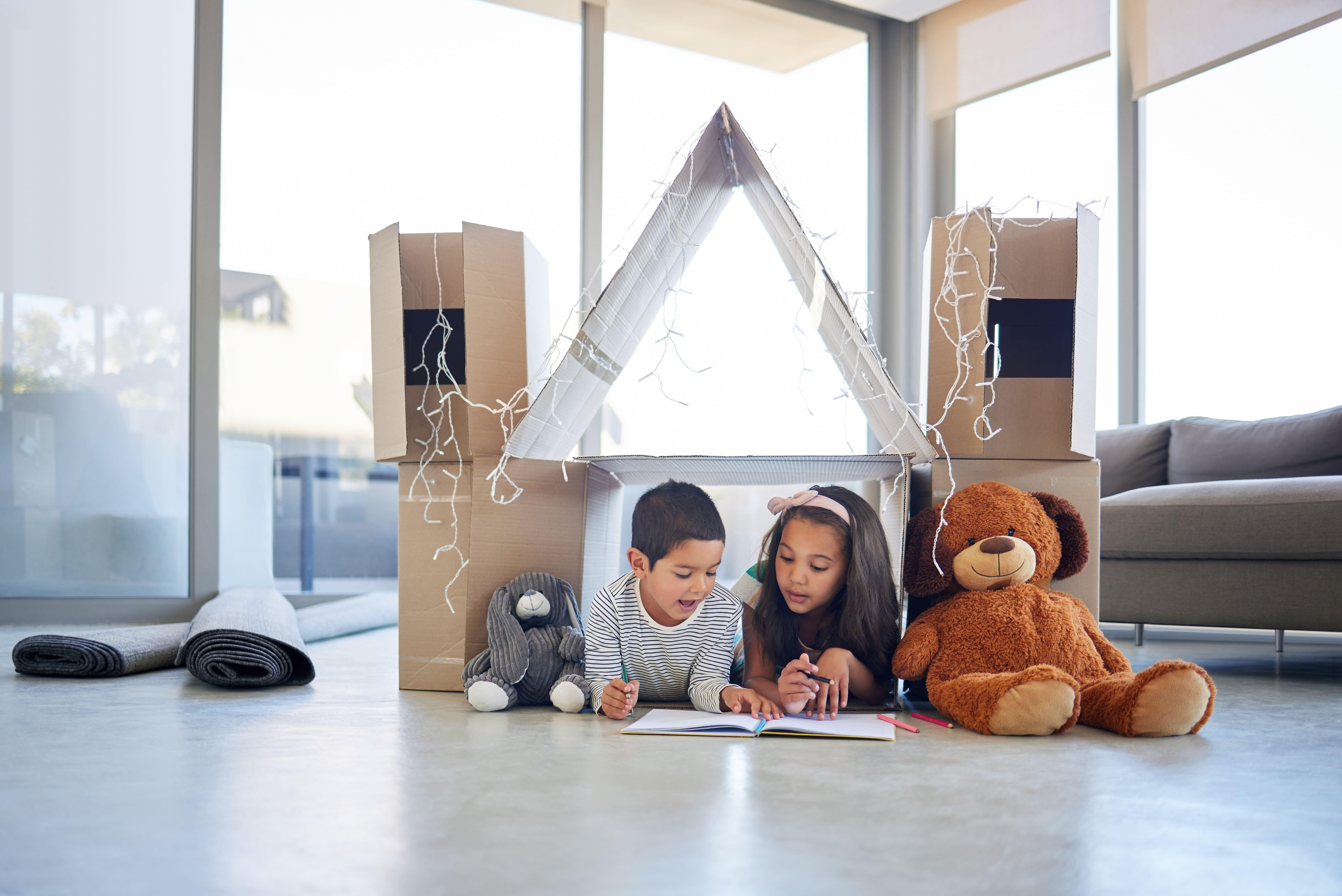 Crianças brincando dentro de uma casa de brinquedo feita com caixas de papelão, cercadas por ursinho de pelúcia e decorada com luzes natalinas, em ambiente ensolarado.
